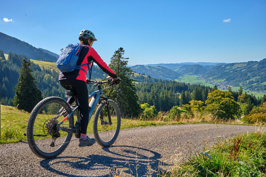 Female mountain biker overlooking a scenic alpine valley with green hills, dense forests and distant villages on a sunny day. Thalkirchdorf, Bavaria, Germany