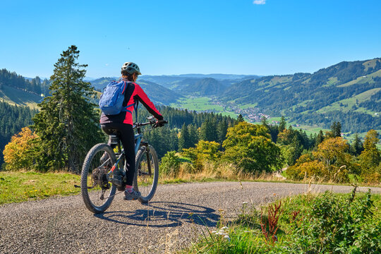 Female mountain biker overlooking a scenic alpine valley with green hills, dense forests and distant villages on a sunny day. Thalkirchdorf, Bavaria, Germany