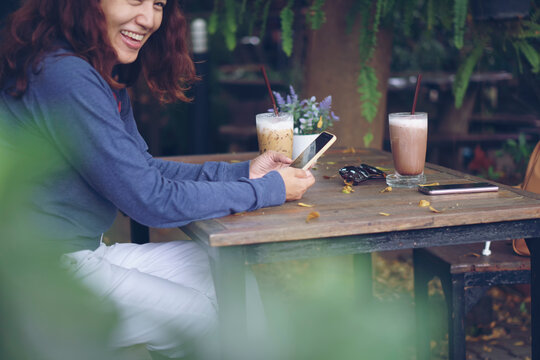 Asian woman smiling using smartphone at outdoor cafe, enjoying coffee and chocolate drink at wooden table with sunglasses and flower. Lifestyle and technology concept.