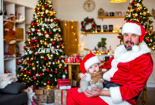 Happy father dressed as Santa Claus holding his baby at home during Christmas, enjoying a warm and joyful family holiday moment.