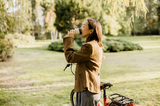 Woman enjoys hot drink while standing next to bicycle in a park during daytime