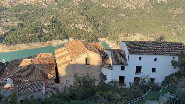 High angle panning shot revealing traditional tile roof houses, turquoise reservoir river, and green mountain landscape during a sunny winter sunset in Montanejos, Spain.