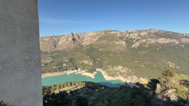 Wide angle static shot showcasing the scenic turquoise reservoir and rugged limestone mountains covered in Mediterranean pine forests under a bright sunny winter sky in Montanejos, Spain.