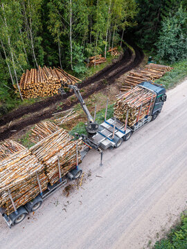 Aerial view of a logging truck with a hydraulic crane loading cut timber logs onto a trailer at a forest site with stacks of wood along a dirt road in Latvia.