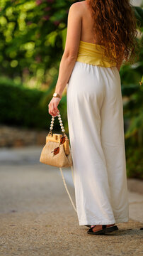 A woman with long curly hair wearing a yellow top and white trousers carrying a straw handbag