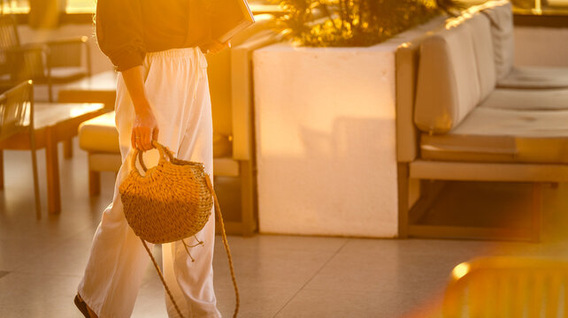 A woman walking through a sunlit outdoor lounge area carrying a stylish woven straw handbag