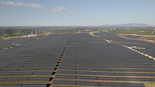 Aerial view of a large solar farm with rows of photovoltaic panels, electrical infrastructure, and rolling hills in the background in Bensafrim, Faro, Portugal.
