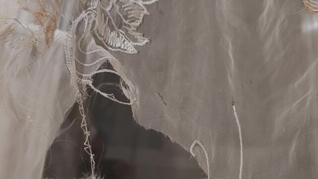 Close up of a weathered, vintage embroidered curtain adorned with delicate floral designs, unveiling a shadowy and enigmatic window. Hinting at the theme of neglect and desolation
