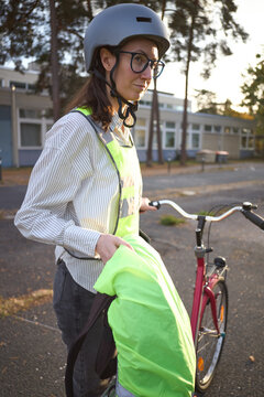 Helmeted woman cyclist holds a fluorescent safety jacket beside her bicycle outdoors