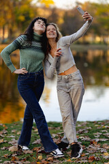 Fototapeta premium Two friends take a cheerful selfie together by a picturesque lake during autumn, surrounded by falling leaves and vibrant colors of the season