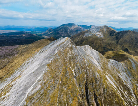 Aerial view of Stob Ban mountain peak in the Mamores range, featuring rocky slopes, patches of grass, and distant ridges under a bright sky in Fort William, Scotland, United Kingdom.