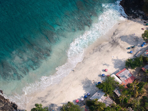 Aerial view of the turquoise waters and white sand beach of Blue Lagoon near Pelabuhan Padang Bai, with colorful umbrellas and lush tropical trees in Kecamatan Manggis, Bali, Indonesia.