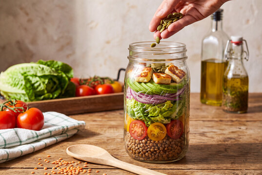 Woman Hand Adding Pumpkin Seeds to Fresh Layered Mason Jar Salad with Halloumi Avocado and Lentils
