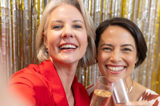 Diverse female friends celebrating, taking selfie in front metallic tinsel with red blouse, flutes