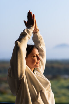 Woman standing on balcony raising arms and pressing palms together, in hoodie and fitness tracker
