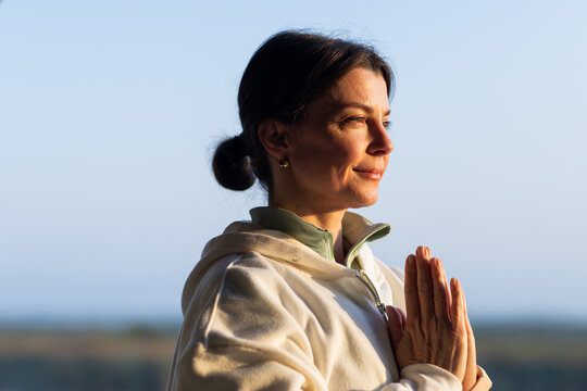 Woman standing with hands together at chest, praying near horizon, wearing hoodie green top hoops