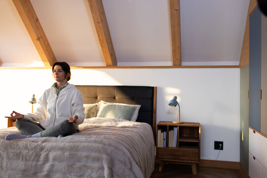 Female meditating on bed in loft bedroom near headboard, bedside lamp, wearing hoodie and leggings