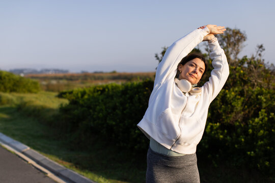 Middle-aged woman stretching on paved path next to curb wearing white hoodie and headphones