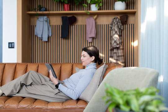 Woman reclining on tan leather sofa at home with light cushion holding tablet, slatted wall plants