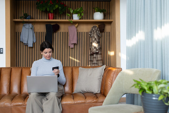 Woman sitting on tan sofa in living room, wearing turtleneck, using laptop, holding credit card