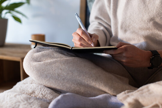 Female writing in black notebook sitting cross-legged at home wearing watch holding white pen
