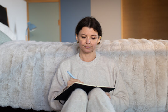 Woman wearing light gray loungewear sitting on floor writing in black notebook with blue pen
