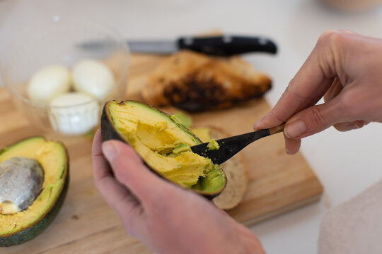 Adult female in light long-sleeve holding avocado half using black spoon on wooden cutting board