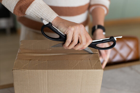Female wearing striped sweater cutting tape with black kitchen shears on cardboard box at table