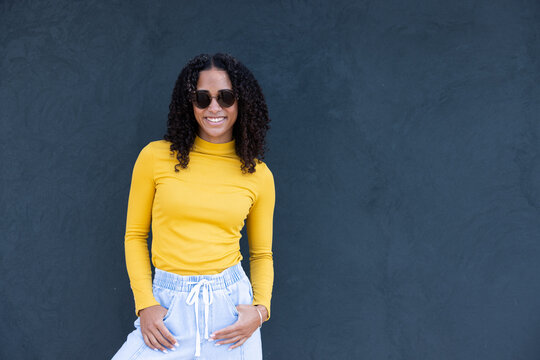 African American woman standing left by slate wall wearing shades, yellow top, bracelet, copy space