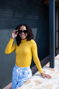 African American woman leaning on stone ledge adjusting sunglasses on walkway wearing yellow top