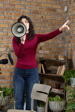 Female adult speaker holding megaphone, pointing, wearing burgundy top and jeans in studio with mic