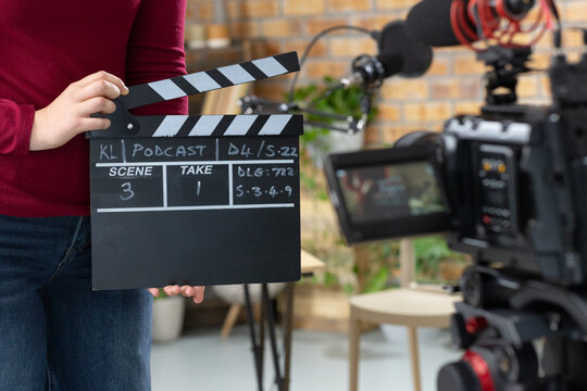 Woman holding clapperboard marked PODCAST, standing on small studio set with camera rig, copy space