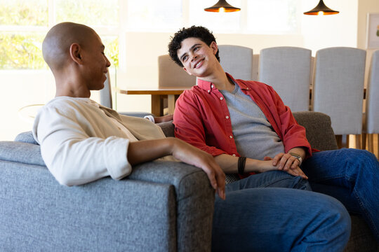 Diverse male friends sitting on gray sofa in open living dining room near large windows, chatting