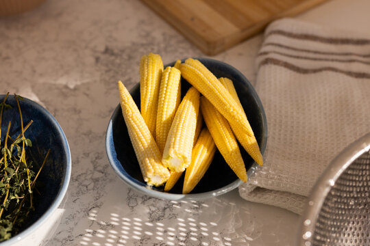 Blue ceramic bowl holding baby corn cobs on marble countertop, with towel and herbs visible