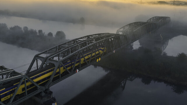 Aerial view of the Spoorbrug Buggenum railway bridge with a passenger train crossing over the river during a misty sunrise in Buggenum, Limburg, Netherlands.