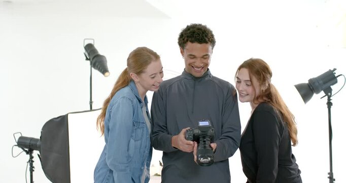 Diverse friends leaning in while male photographer scrolling DSLR, reviewing shots in white studio