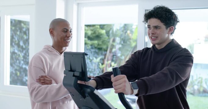 African American men on elliptical at home, rider pedaling, partner seeing strain adjusting console