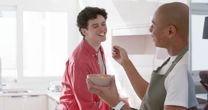 African American man in apron with bowl offering carrots urging teen male leaning cooking at stove