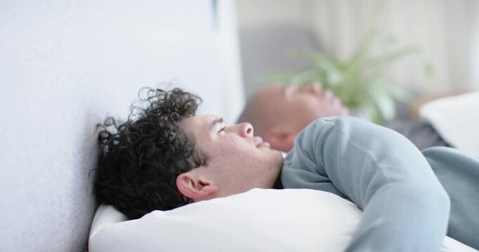 Two men, curly man pressing ear against white pillow, rubbing head, reacting to noise in bedroom