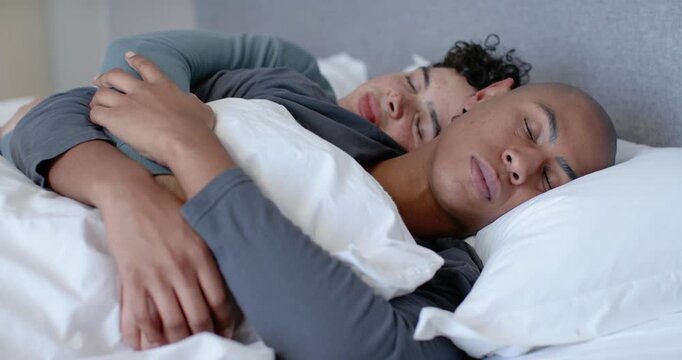 Two men lying spooning under white duvet in bedroom, rear man adjusting grip seeking comfort