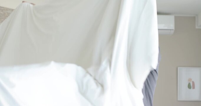 African American man lifting and shaking white duvet to fluff and straighten bed in bedroom