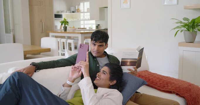 Diverse couple on couch, woman scrolling pink phone, urging man leaning over booklet to view