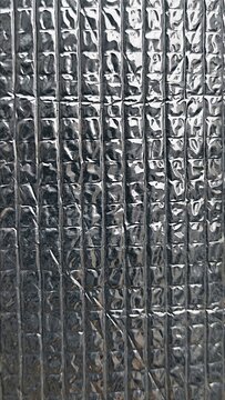 ​A vertical close-up shot of a silver foil thermal insulation sheet featuring a embossed grid pattern with shiny light highlights and dark metallic reflections