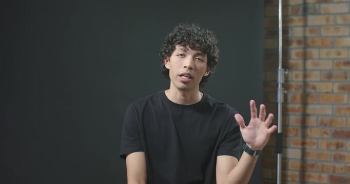 African American man facing camera, sitting on set by brick wall in black tee, gesturing