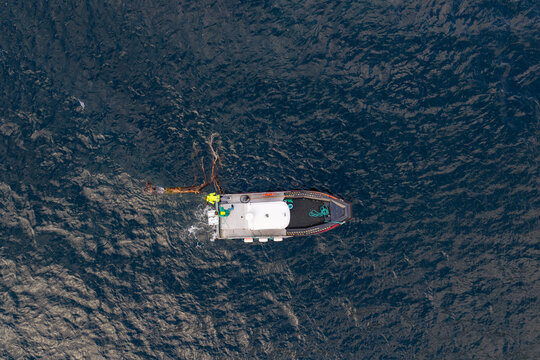 Aerial view of a workboat towing a large tree away from a fish farm in the deep blue waters of a fjord in Fjord, More og Romsdal, Norway.