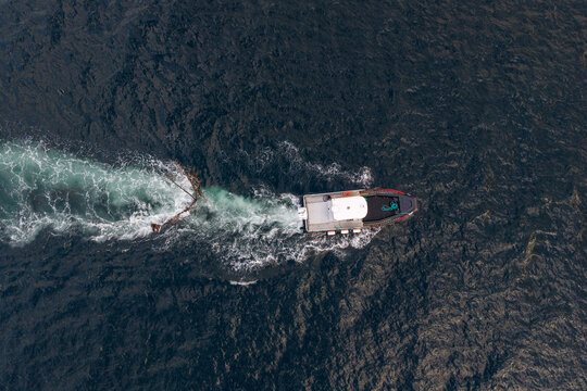 Aerial view of a workboat towing a large tree away from a fish farm in the dark waters of a fjord in Fjord, More og Romsdal, Norway.