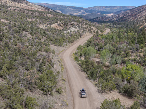 Aerial view of a teardrop camper trailer traveling overland along the dirt road of Hells Backbone surrounded by rugged mountains and pine trees in Garfield County, Utah, United States.