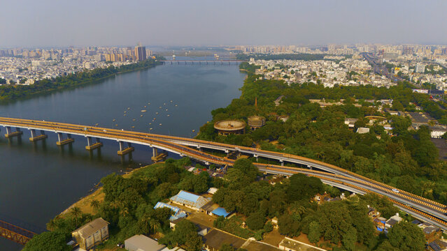 Aerial view of the Tapi River flowing through the urban landscape of Surat, featuring a prominent bridge, lush green vegetation, and dense city buildings in Surat, Gujarat, India.