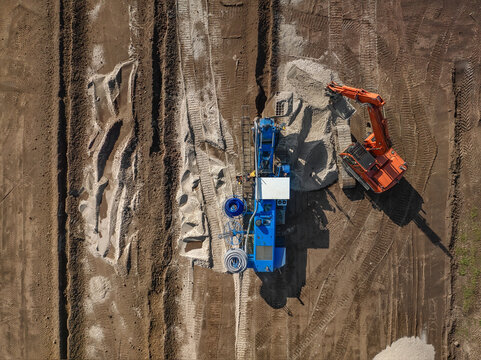 Aerial view of construction machinery including an orange excavator and a blue specialized drilling rig working on a large brown soil site in Enschede, Overijssel, Netherlands.