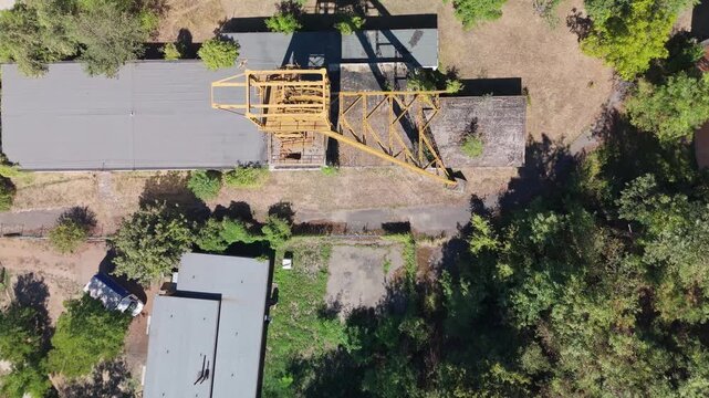Top-down drone view of a former coal mine headframe, industrial buildings and overgrown grounds amid surrounding summer trees in B&aacute;tonyterenye, Hungary.